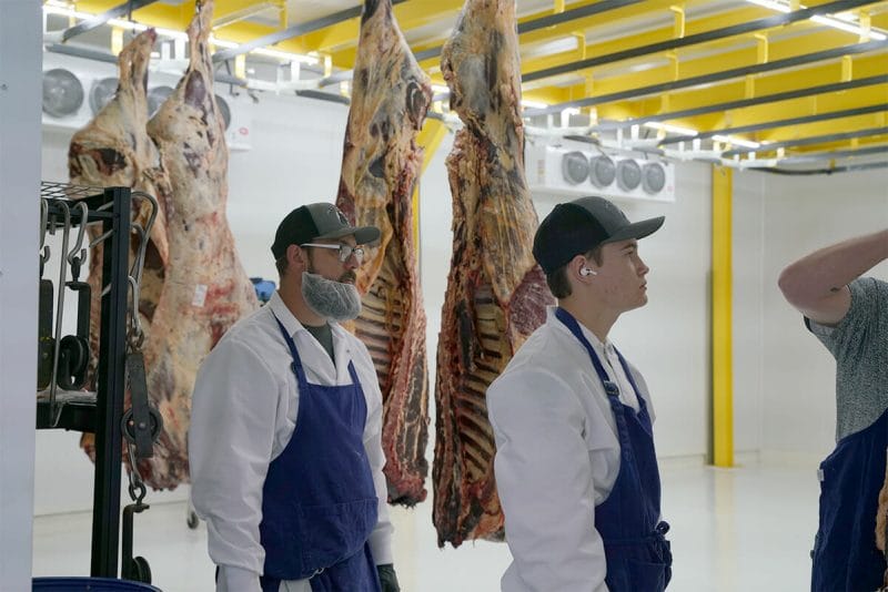 Two men standing in the walking cooler at 160 Processing looking at another man working on some of the meat that is hanging.